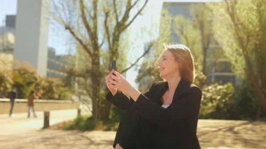 Uzun saçlı ve mavi gözlü genç bir iş kadını modern bir kentsel alanda bankta oturuyor, akıllı telefonuyla yaptığı bir telefon görüşmesinde gülümsüyor ve gülüyor..