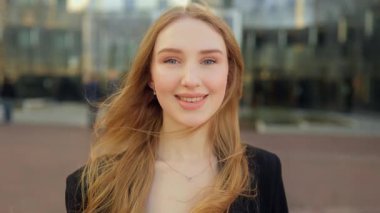 Dışarıda duran uzun saçlı çekici genç iş kadını, kameraya sıcak bir şekilde gülümserken gevşek saçlarını doğal ve kendinden emin şehir portresini düzeltiyor..