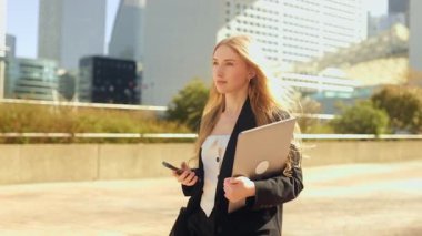 Kendine güvenen, uzun saçlı genç bir iş kadını elinde akıllı bir telefon ve dizüstü bilgisayarla modern bir caddede yürüyor. Odaklanmış ve kararlı bir şehir kurumsal yaşam tarzı var..