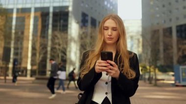 Takım elbiseli genç bir iş kadını açık havada, büyük bir şehirde akıllı telefon kullanarak yürüyor. Güzel bayan profesyonel yönetici cep telefonunda yazıyor, çevrimiçi mobil teknoloji iş uygulamaları