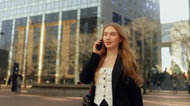 Resmi takım elbiseli, gülümseyen, uzun saçlı bir kadın cep telefonuyla konuşuyor, Paris 'teki bir iş merkezinin caddesinde yürüyor, akıllı telefon, yavaş çekim, cam binaların arka planında.