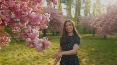Genç bir kadın çiçek açan bir sakura bahçesinde yürüyor, kiraz çiçeklerine hafifçe dokunuyor ve kameraya gülümsüyor. Romantik bahar anı, yumuşak ışık, çiçekli güzellik, zarafet ve doğal zarafet..