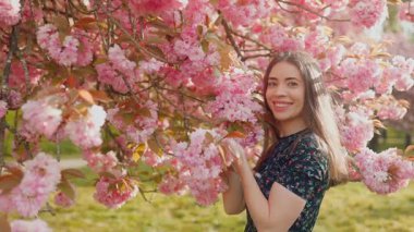 Çiçek açan bir sakura dalının yanında poz veren güzel genç bir kadın, çiçekleri nazikçe kokluyor ve kameraya gülümsüyor. Göz kamaştırıcı arka plan, romantik bahar atmosferi, pembe çiçekler, yumuşak güneş ışığı, zarif ve kadınsı ruh hali..