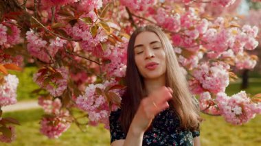 Genç ve çekici bir kadın çiçek açan bir sakura bahçesinde kameraya öpücük yolluyor. Romantik bahar anı, güzellik ve doğadaki olumlu duygular..