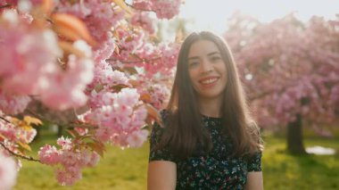 Genç ve çekici bir kadın kameraya gülümsüyor ve çiçek açan bir sakura bahçesinde ellerinden kiraz çiçekleri saçıyor. İlkbahar güzelliği, neşe ve doğa konsepti