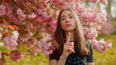 Genç bir kadın neşeyle başını sallıyor ve parmağıyla bir jest yapmıyor. Hoş bir şekilde onaylamadığını ifade ediyor. Sonra da sakura ağacının altında içten bir gülümseme sergiliyor.