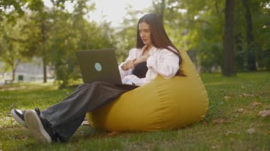 Genç kadın puf minderinin üzerine oturmuş video ile iletişim kuruyor. Laptop bilgisayarına bakan bir kız, online konferans ofis sohbeti, dışarıda çalışan sanal eğitim konsepti bilişim uzmanı. Yüksek kalite 4k görüntü
