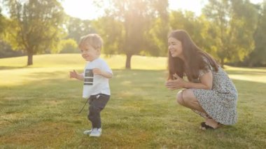 Anne yanında yürürken ve destek için alkışlarken mutlu çocuk ilk adımlarını atıyor. Duygusal dönüm noktası, neşeli çocukluk anı, bağlanma, teşvik, güneşli doğada yürümeyi öğrenme.