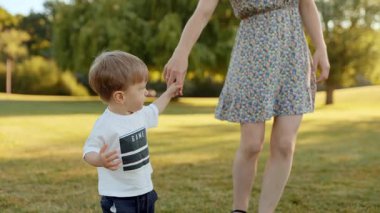 Yürüyen çocuk güneşli bir çayırda annenle el ele yürüyor. Tatlı aile anı, kaynaşma, yürümeyi öğrenme, aşk, destek, çocukluk keşfi, doğa, yaz ışığı, duygusal sahne.