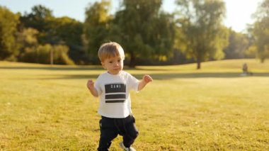 Mutlu çocuk parktaki çimenli çayıra ilk adımını atıyor, gururla gülümsüyor. Bebek yürüyüşü, kilometre taşı anı, çocuk gelişimi, doğa geçmişi, aile hayatı sahnesi.