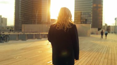 İş kadını güvenli bir şekilde ofis binasına doğru yürüyor, arkadan görülüyor. Gün batımı ışığı onun siluetini özetliyor. Güç, hırs ve amaç. Her adımda modern liderlik harekete geçiyor..