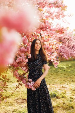 Geniş bir sakura bahçesinin önünde duran genç bir kadın, arkasında kiraz çiçekleri olan dikey bir portre, yumuşak bir şekilde gülümsüyor, rüya gibi bahar zamanı güzelliği..