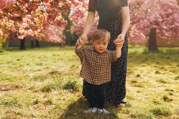 Annesi çiçek açan bir kiraz bahçesinde elini tutarken kameraya gülümseyen bir çocuk. Bahar doğasında neşeli bir aile anı.