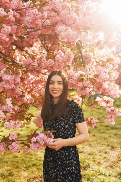 Açık havada duran gülümseyen genç bir kadın, doğal ışıkla dikey bir portre, neşeli bir ifadeyle kameraya bakıyor, çiçek açan sakura arka planlı bahar güzelliği.