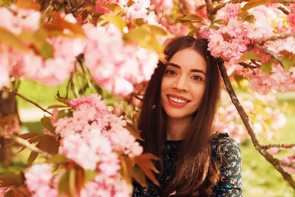 Güzel genç bir kadın çiçek açan kiraz çiçeklerinin altında kameraya gülümsüyor, sakura çiçekleriyle bahar portresi, doğal ışık, neşeli ruh hali, mevsimsel güzellik, yatay fotoğraf.