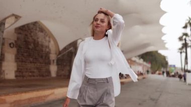 Paris 'te Seine nehrinde yürüyen, saçları düzelten, gülümseyen, şehir manzarasına hayran olan uzun saçlı mutlu genç kadın kameraya dönüyor. Seyahat, yaşam tarzı, moda, romantik şehir manzarası.