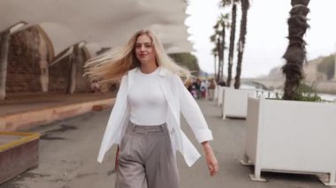 Paris 'te Seine nehri kıyısında yürüyen uzun saçlı, gülümseyen genç bir kadın, kameraya dönüyor, neşeli bir ifade. Seyahat, yaşam tarzı, şehir manzarası, moda, mutluluk.