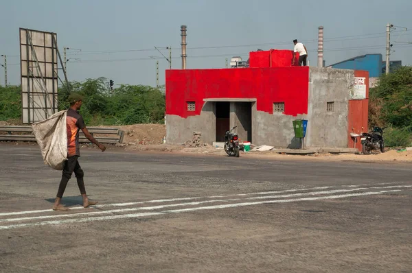 Indian young man crosses the road with a sack. Another man is painting the house red. Two bicycles are parked near the house.