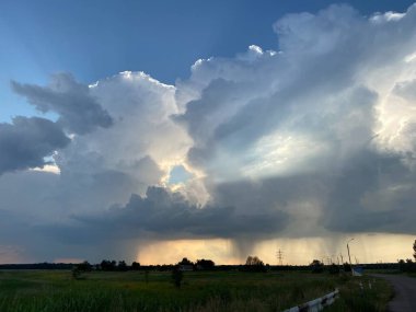 Thunderstorm clouds and somewhere far away it rains against the blue sky and trees and a green field on the horizon.