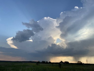 Thunderstorm clouds and somewhere far away it rains against the blue sky and trees and a green field on the horizon.