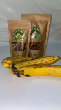 Beyaz bir masada kurutulmuş muz cipsi ve kese kağıdının yanında olgun sarı muzlar..