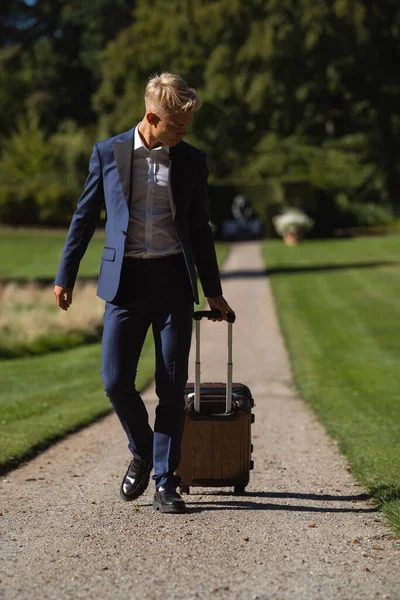 A man in a blue suit walks with a suitcase on a gravel path