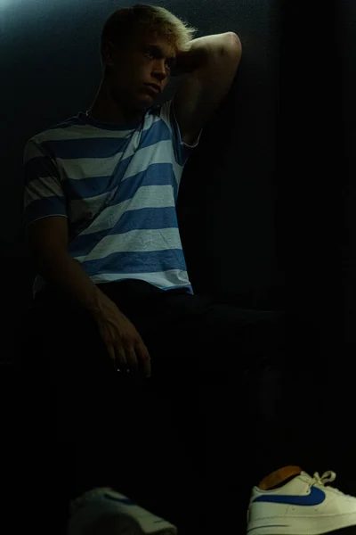 A thinking man in a striped T-shirt sits in a dark room with a soft natural light falling on him