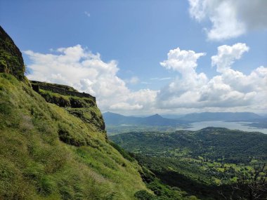 Fotoğraf, gür yapraklarla kaplı ve kır çiçekleriyle süslenmiş bulutlarla dolu kayalık bir arazinin büyüleyici bir görüntüsünü tasvir ediyor..
