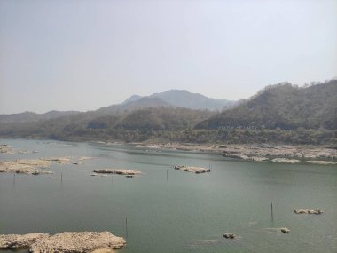 Görüntü, dağlarla çevrili büyük bir su kütlesinin ve berrak bir gökyüzünün bulunduğu huzurlu bir manzarayı tasvir ediyor..