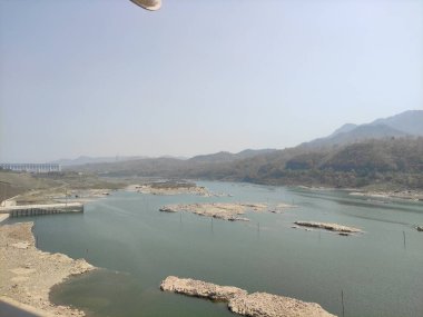 Resim, geniş bir su kütlesi, arka planda dağlar ve açık bir gökyüzü olan sakin bir çevreyi gösteriyor..