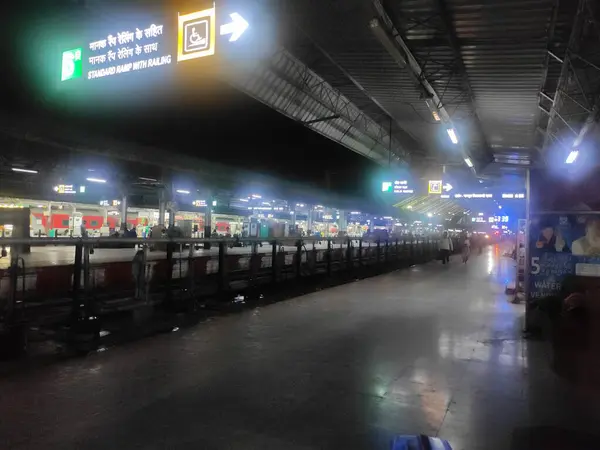 Görüntü, geceleri hareketli bir tren istasyonunu, banklarla kaplı uzun bir platformu ve demiryolu ile standart rampanın yönünü gösteren bir tabelayı tasvir ediyor..