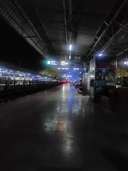 Resim, geceleri yoğun bir tren istasyonunu, uzun bir platformu kaplayan bankları ve demiryolu tepesindeki tipik rampayı gösteren bir tabelayı gösteriyor..