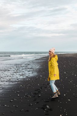 Blond Woman in Yellow Coat Walking on Beach with Black Sand, Diamond Beach in Iceland, Atlantic Ocean  