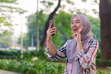 Gülümseyen Asyalı kadın parkın açıklarında akıllı telefonuyla video görüşmesi yapıyor.