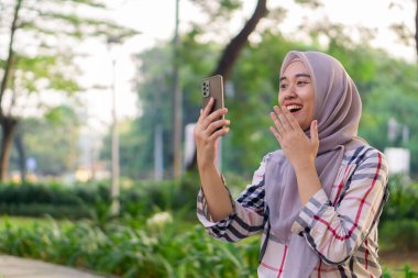 Gülümseyen Asyalı tesettürcü kız dışarı çıktı ve parkta akıllı telefon oynarken şaşırdı.