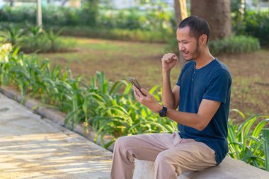 Mavi gömlekli genç Asyalı adamlar parkta akıllı telefon çalarken gülümsüyor ve gülüyorlar.