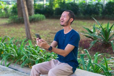 Mavi gömlekli genç Asyalı adamlar parkta akıllı telefon çalarken gülümsüyor ve gülüyorlar.