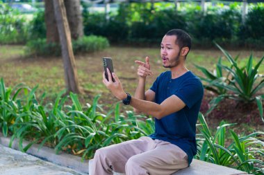 Mavi gömlekli genç Asyalı adamlar parkta akıllı telefon çalarken gülümsüyor ve gülüyorlar.