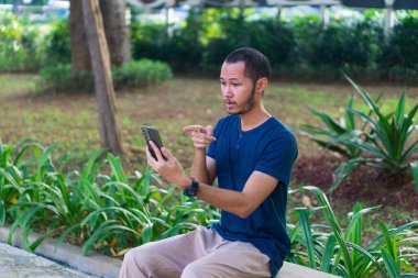 Mavi gömlekli genç Asyalı erkekler parkın dışında akıllı telefon çalarken şok geçirip strese giriyorlar.