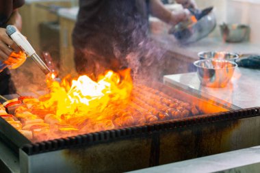 Şefin ellerini mangalda sosis şiş pişirirken, karides ve sebzeler ızgarada cızırdarken yakın çekim. Ateşle