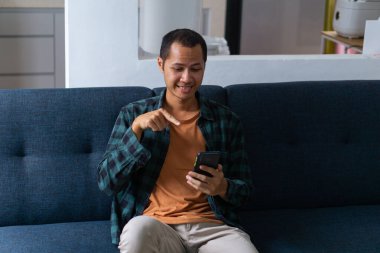 Turuncu tişört ve flanel giyen genç adam koltuktaki akıllı telefonu işaret ediyor..