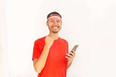 Yumruğunu sıkmış genç Endonezyalı bir adam. Elinde saç bandıyla, izole edilmiş beyaz bir cep telefonu tutuyor.