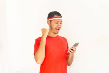 Yumruğunu sıkmış genç Endonezyalı bir adam. Elinde saç bandıyla, izole edilmiş beyaz bir cep telefonu tutuyor.