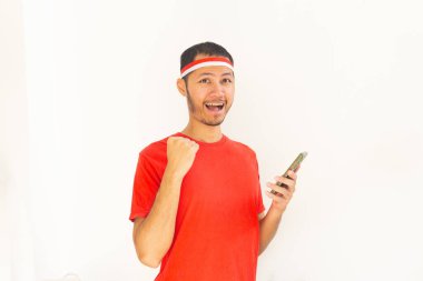 Yumruğunu sıkmış genç Endonezyalı bir adam. Elinde saç bandıyla, izole edilmiş beyaz bir cep telefonu tutuyor.