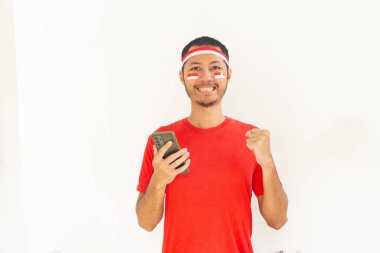 Yumruğunu sıkmış genç Endonezyalı bir adam. Elinde saç bandıyla, izole edilmiş beyaz bir cep telefonu tutuyor.