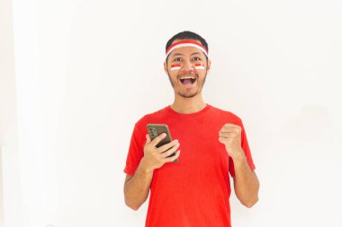 Yumruğunu sıkmış genç Endonezyalı bir adam. Elinde saç bandıyla, izole edilmiş beyaz bir cep telefonu tutuyor.