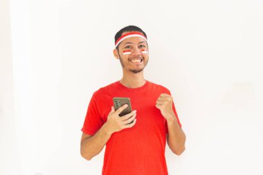 Yumruğunu sıkmış genç Endonezyalı bir adam. Elinde saç bandıyla, izole edilmiş beyaz bir cep telefonu tutuyor.