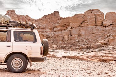 Rear side view of a 4x4 car loading package and wood in the Sahara desert of Tadrart Rouge. Parked in the sandstone reg with in the Sahara desert, rocky mountain with cloudy sky in the background.