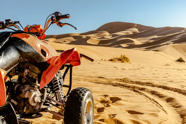 Quad bike orange and black color vehicle parked  in the Sahara desert, selective focus in foreground, golden yellow colored sand dunes and blue sky in blur background. Rear view of the front side part