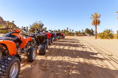 Taghit, Bechar, Cezayir - 27 Aralık 2022: Turistler tarafından kiralanmaya hazır park edilmiş kuadlar. Güneşli bir gün. Kum, palmiye ağaçları ve kuyruktaki bisikletlerin yanında duran tanınmayan turistler..